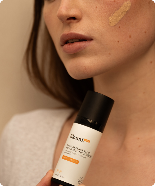 Een close-up van een persoon met een kleine veeg van Likami Daily Defence Fluid 'Correct + Brighten' op hun wang, terwijl ze de bijbehorende fles vasthouden.