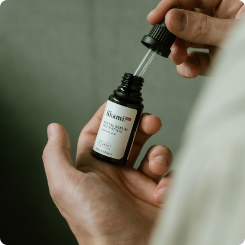 Een close-up van handen die een geopend donker glazen flesje van het Likami Hydration serum vasthouden en de pipet eruit trekken. De afbeelding heeft afgeronde hoeken.