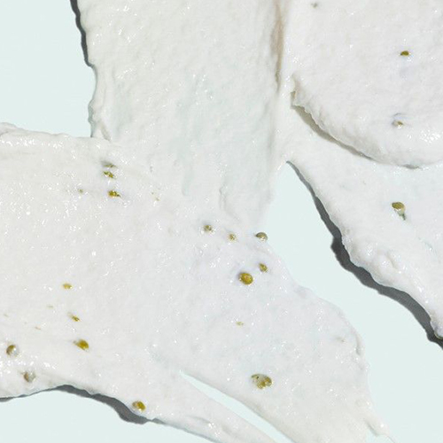 Een gedetailleerde weergave van de romige, witte textuur van de Iluma cleanser met zichtbare kleine, blauwe scrubdeeltjes.