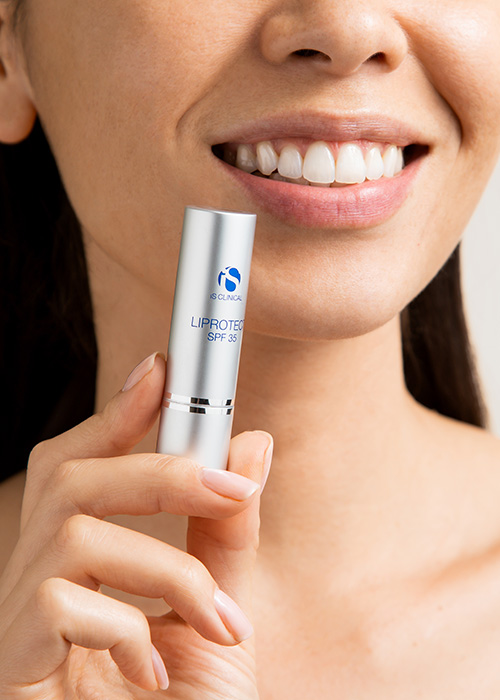 Een lachende vrouw houdt een zilveren huls van iS CLINICAL Liprotect SPF 35 lippenbalsem voor haar gezicht.