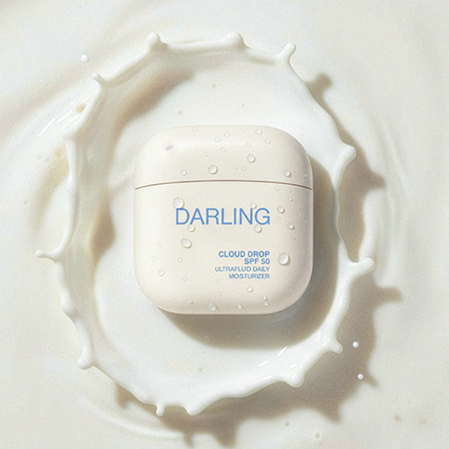 De Darling Cloud Drop flacon ligt in het midden van een vloeibare, melkachtige witte textuur met spatten.