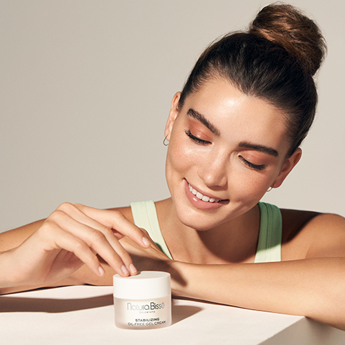 Een vrouw kijkt glimlachend naar het potje Natura Bissé Stabilizing Oil-Free Gel Cream dat voor haar op tafel staat.