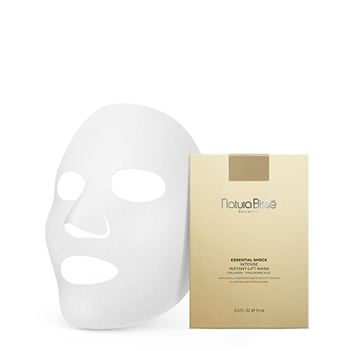 Een wit bladmasker naast een goudkleurige verpakking van Natura Bissé Essential Shock Intense Instant Lift Mask. De verpakking toont de merknaam en productdetails. De achtergrond is wit.