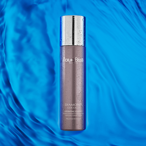 De Hydrating Essence flacon van Natura Bissé met waterdruppels, liggend op een golvende blauwe achtergrond die doet denken aan water.