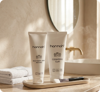Een vierkante close-up weergave, perfect voor mobiel, van de hannah 'Enzymatic Skin Peeling' en 'Cleansing Clay' tubes, geplaatst op een beige plateau met een applicatiekwast in een badkameromgeving met een ronde spiegel.