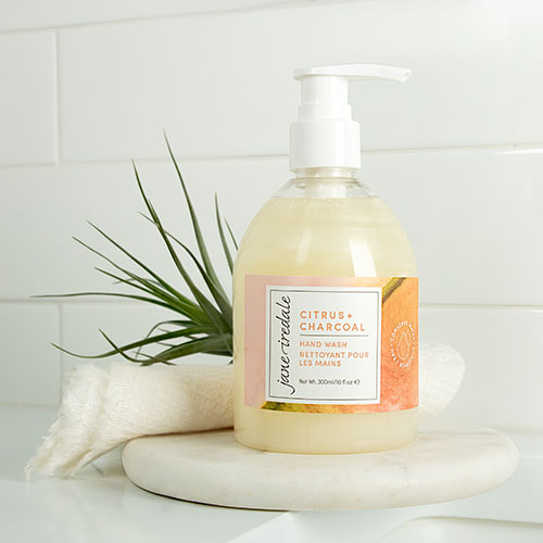 Jane Iredale Citrus + Charcoal Hand Wash staat op een wit marmeren plateau in een badkamerinterieur. Naast de pompflacon ligt een opgerolde handdoek met een groen luchtplantje.