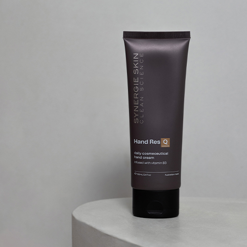 Een sfeerfoto van de staande tube Synergie Skin Hand ResQ handcrème op een lichtgrijs stenen plateau tegen een egale grijze achtergrond.