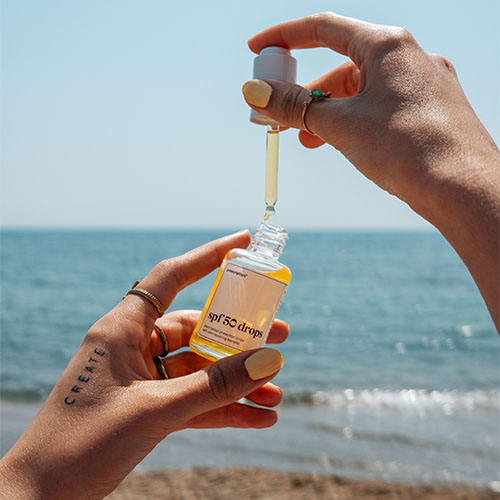 Een persoon houdt een flesje Prescription spf 50 drops boven een ander flesje en laat er serum in druppelen. De achtergrond is de zee en het strand. De nagels zijn geel gelakt.