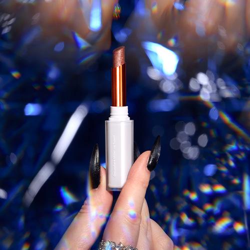 Hand met opvallende nagels presenteert de open taupekleurige Nimya glitter lippenstift voor een glinsterende blauwe achtergrond