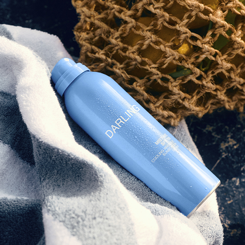 Een lichtblauwe spuitbus van Darling Mist Me SPF 50 ligt op een wit met grijs gestreepte handdoek. Op de achtergrond is een rieten mand gevuld met citroenen zichtbaar onder het warme zonlicht.