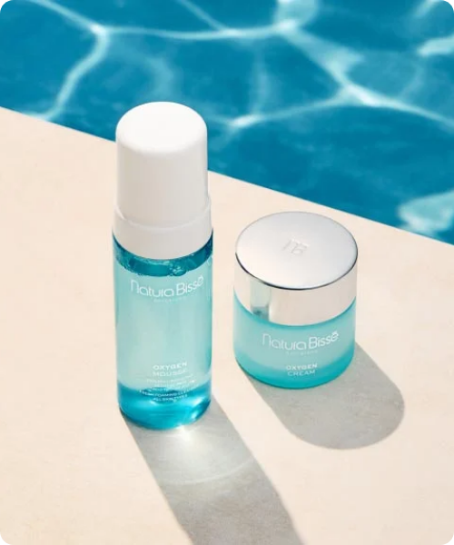 Een flacon Natura Bissé Oxygen Mousse en een potje Oxygen Cream staan naast elkaar op een beige rand bij een helderblauw zwembad. De afbeelding heeft afgeronde hoeken.