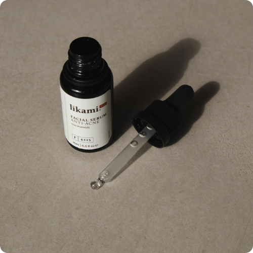 Een glazen flesje van het Likami Anti-Acne serum met de gevulde pipet ernaast, weerspiegeld op een glanzende witte ondergrond. De afbeelding heeft afgeronde hoeken.