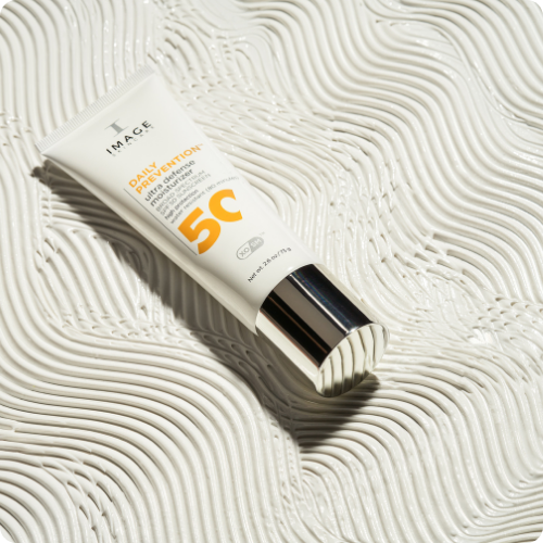 Een tube IMAGE Skincare Daily Prevention Ultra Defense Moisturizer SPF 50 ligt schuin op een ondergrond van witte, golvende crèmetextuur.