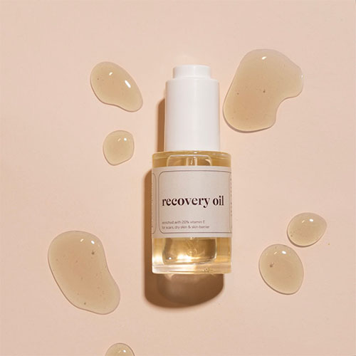 Een platte foto van een flesje Prescription recovery oil met witte dop, omringd door gemorste druppels olie op een roze achtergrond. De tekst op het label geeft aan dat het product 20% vitamine E bevat.