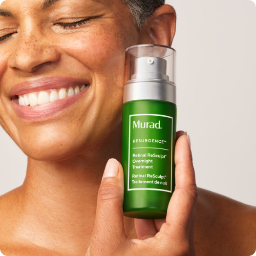Een vrouw toont lachend een groene flacon Murad Retinal Resculpt Overnight Treatment tegen haar gezicht