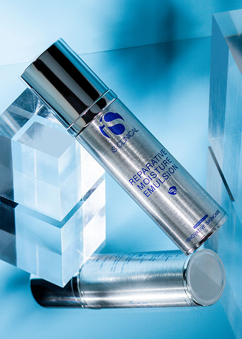 Een glanzende flacon iS CLINICAL Reparative Moisture Emulsion staat schuin op een lichtblauwe ondergrond, omringd door transparante kubussen, met een tweede flacon die eronder op de ondergrond ligt.