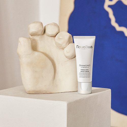 Een tube Natura Bissé Diamond Luminous Perfecting Hand Cream staat op een witte sculpturale kubus, naast een beige stenen beeldhouwwerk van een hand, tegen een achtergrond met blauwe vormen.