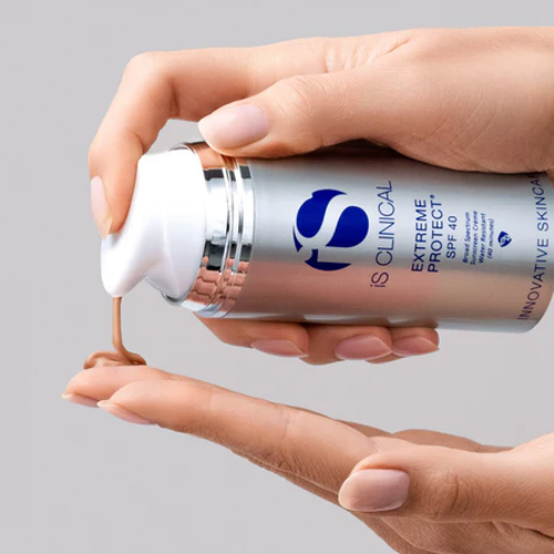 Hand die een getinte, bruingekleurde zonnebrandcrème pompt uit de zilveren flacon van iS Clinical Extreme Protect SPF 40 op de vingers van een andere hand tegen een grijze achtergrond
