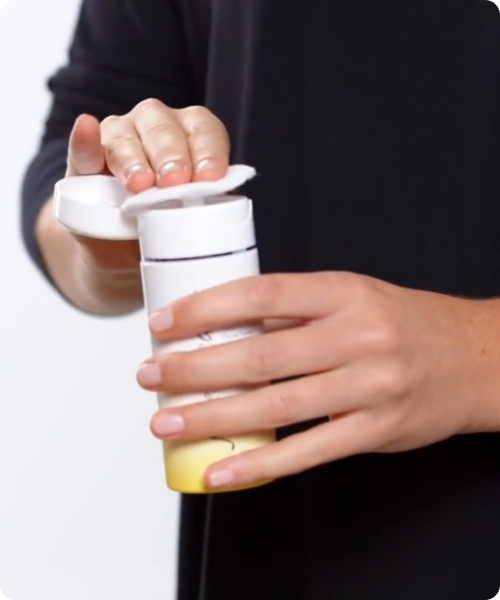 Close-up van handen die een wattenschijfje op de doseerpomp van een Jane Iredale BeautyPrep Toner drukken. De flacon heeft een wit-naar-geel kleurverloop.