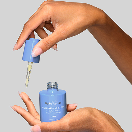 Handen die de blauwe flacon en pipet van de HydroPeptide Micro Dose Glow Booster vasthouden tegen een egale achtergrond.