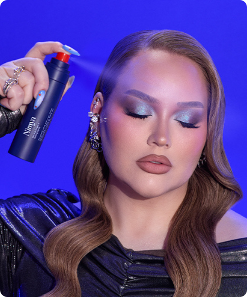 Nikkie de Jager sprayt Nimya setting spray over haar gezicht met gesloten ogen, voor een felblauwe achtergrond met glamour make-up en blauwe oogschaduw.