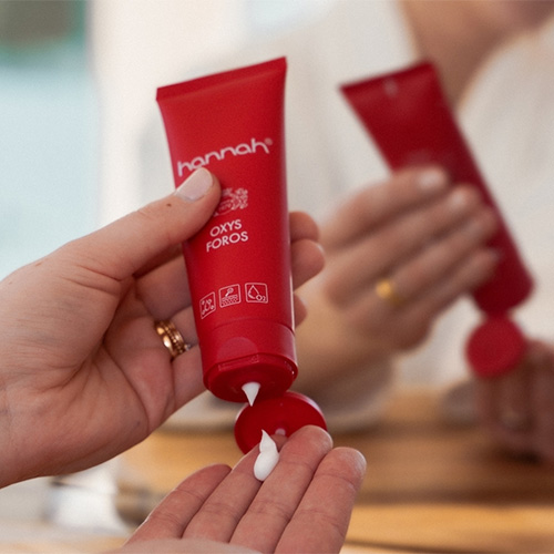 Een close-up van handen die witte crème uit een rode tube van hannah oxys foros knijpen op een handpalm, met op de achtergrond een onscherpe reflectie in de spiegel.