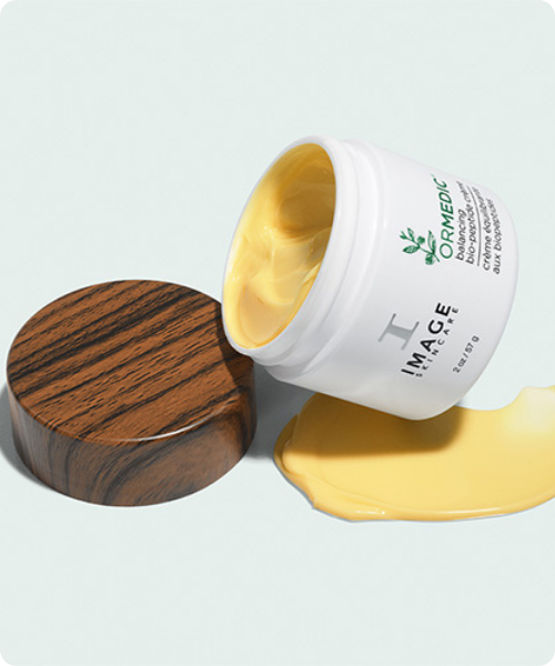 Een geopende pot IMAGE Skincare ORMEDIC Balancing Bio-Peptide Cream met een rijke, gele textuur die naast de pot is uitgestreken op een lichtblauwe achtergrond.