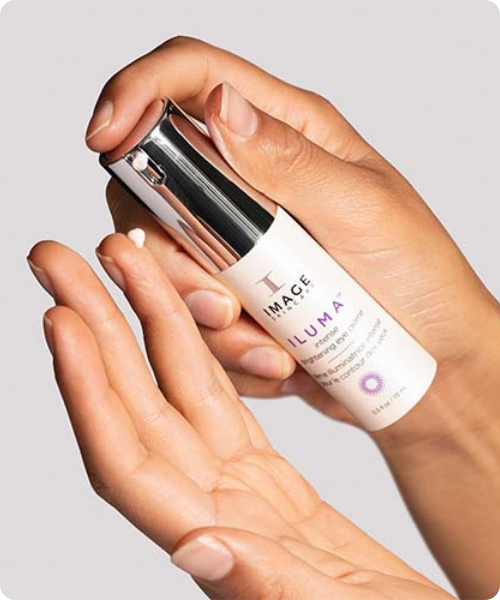 Twee handen die een witte flacon van de ILUMA oogcrème vasthouden, waarbij een kleine druppel crème op een vingertop wordt gedoseerd.