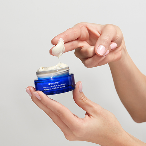 Handen die een geopende blauwe pot HydroPeptide Power Lift vasthouden met rijke crème op de vinger.