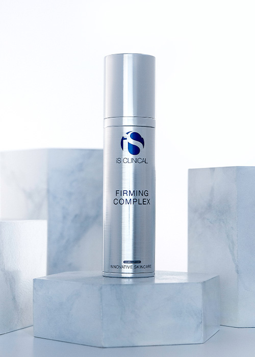 Een flesje iS Clinical Firming Complex staand op een marmerachtig blok, omringd door soortgelijke blokken tegen een lichte achtergrond.