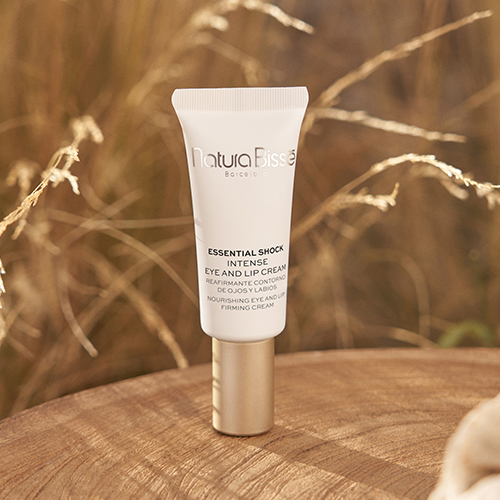 Een witte tube Natura Bissé Essential Shock Intense Eye and Lip Cream met een gouden dop, rechtopstaand op een houten oppervlak tegen een wazige achtergrond van droog gras. De verpakking toont de merknaam en productdetails.