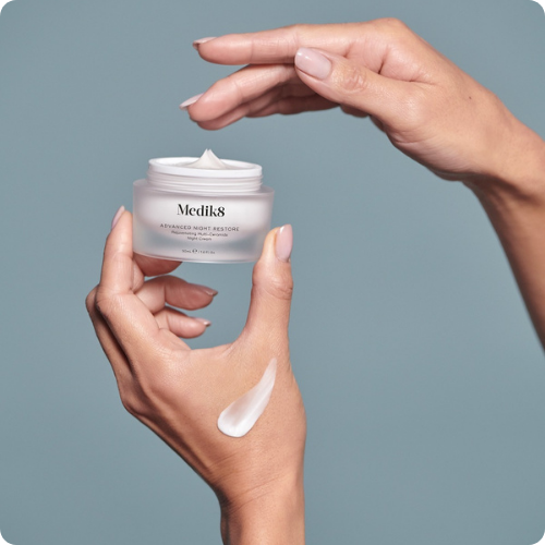 Twee handen houden een geopend potje Medik8 Advanced Night Restore vast met een veeg witte crème op de rug van de hand tegen een blauwgrijze achtergrond.