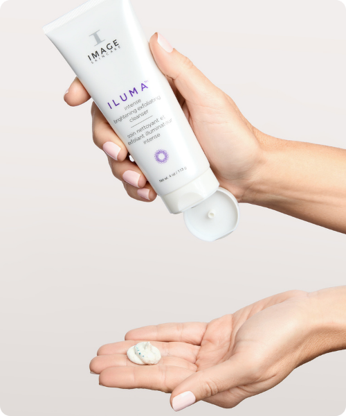 Een hand die de witte tube van de IMAGE Skincare ILUMA exfoliating cleanser vasthoudt en een kleine hoeveelheid crème met blauwe korreltjes in de palm van de andere hand aanbrengt.