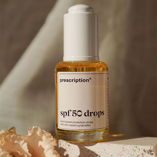 Een close up van een flesje Prescription spf 50 drops op een beige ondergrond, naast een bloemblaadje. Het zonlicht schijnt op het flesje. Het label geeft aan dat het een plant based product is.