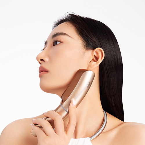 Een vrouw in zijaanzicht die het goudkleurige JOVS Slimax anti-aging apparaat zachtjes tegen haar kaaklijn en hals houdt voor een liftende huidbehandeling