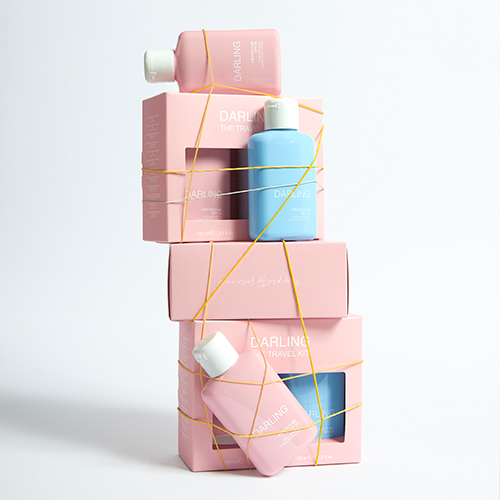 Meerdere roze Darling Travel Kits en losse flacons op een witte achtergrond, speels bij elkaar gehouden door gele elastieken.