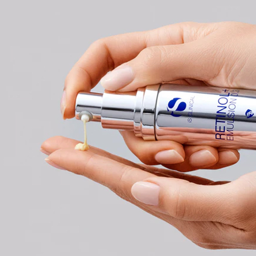 Handen die een gelige crème pompen uit de zilveren flacon van iS Clinical Retinol+ Emulsion 0.3 in de handpalm