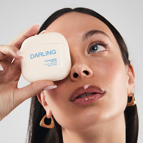 Een model houdt de beige Darling Cloud Drop flacon voor haar oog terwijl ze omhoog kijkt.