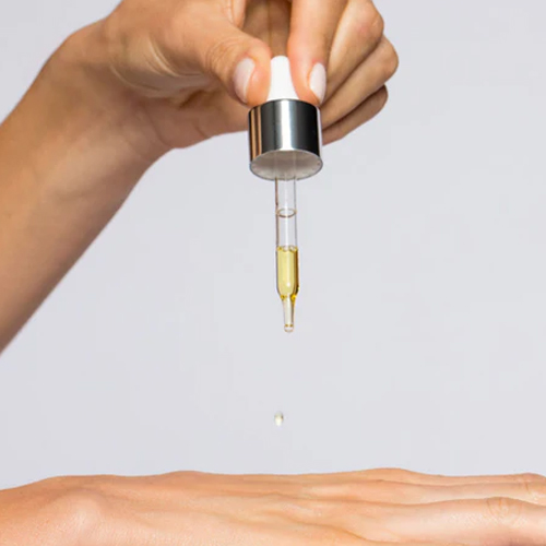 Iemand houdt een glazen druppelaar vast die een goudkleurige druppel serum boven een hand laat vallen.