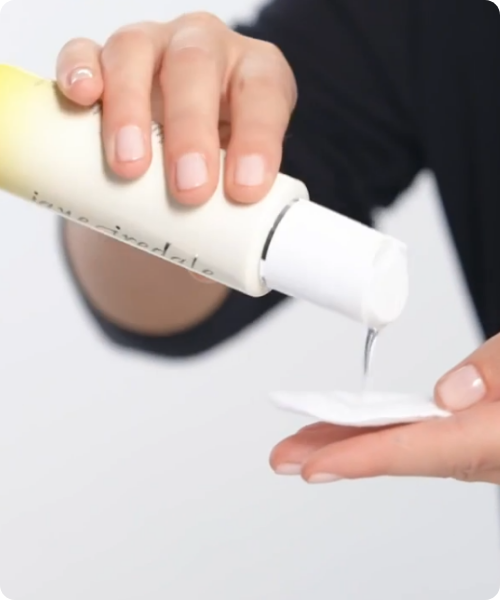 Handen die een doorzichtige reiniger uit een Jane Iredale BeautyPrep Cleanser flacon op een wit wattenschijfje gieten.