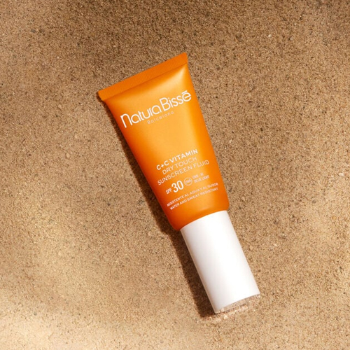 Een oranje tube Natura Bissé C+C Vitamin zonnebrandvloeistof SPF 30 die plat in het zand ligt onder warm, natuurlijk zonlicht.