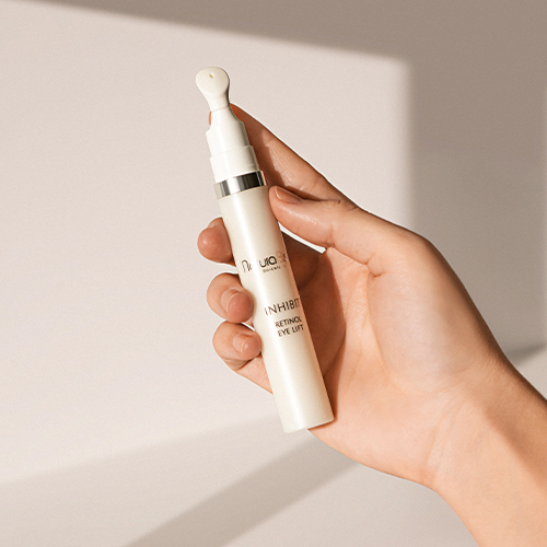 Een hand houdt de Inhibit Retinol Eye Lift tube vast tegen een beige achtergrond met natuurlijke lichtinval.