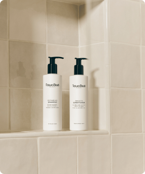 Twee witte flacons met zwarte pompjes, de Natura Bissé Repairing Shampoo en Smooth Conditioner, staan naast elkaar op een tegelplank in een douche-nis.