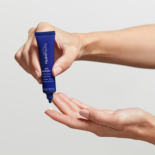 Iemand houdt een blauwe tube HydroPeptide Eye Authority oogcrème vast en brengt een kleine hoeveelheid crème aan op de handpalm.